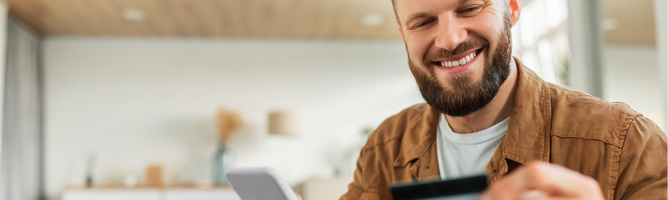 Young adult man smiling while holding mobile phone and looking at his credit card